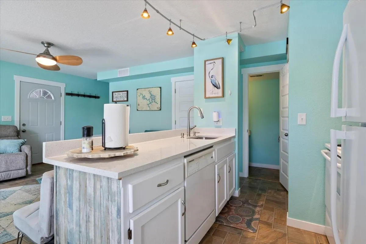 A view of the kitchen with a prominent island, stylish light fixtures overhead, turquoise accent walls, and white cabinetry. The space looks bright and welcoming, designed for both culinary creativity and casual conversation.