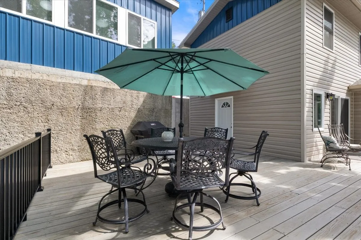 An inviting outdoor dining area with a table, metal chairs, umbrella, and a grill visible in the background. The setup on the patio or deck suggests a perfect spot for grilling meals and gathering with friends or family while enjoying the fresh air and scenic surroundings.