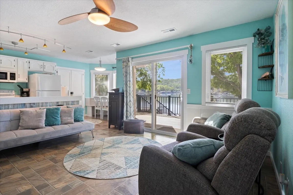 This image captures an open-plan living and dining area featuring a comfortable sofa, a spacious dining table, a decorative rug, and large windows that offer a clear outdoor view of the lake. The setup is ideal for socializing, relaxing, and dining together, while the scenic backdrop connects the interior to the natural surroundings.