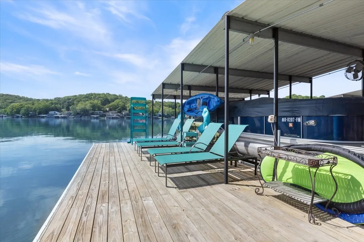 This image captures a tranquil exterior scene featuring a dock with lounge chairs and a kayak resting nearby. The water is calm and inviting, perfect for swimming or kayaking, and the overall setting is peaceful, surrounded by lush greenery.