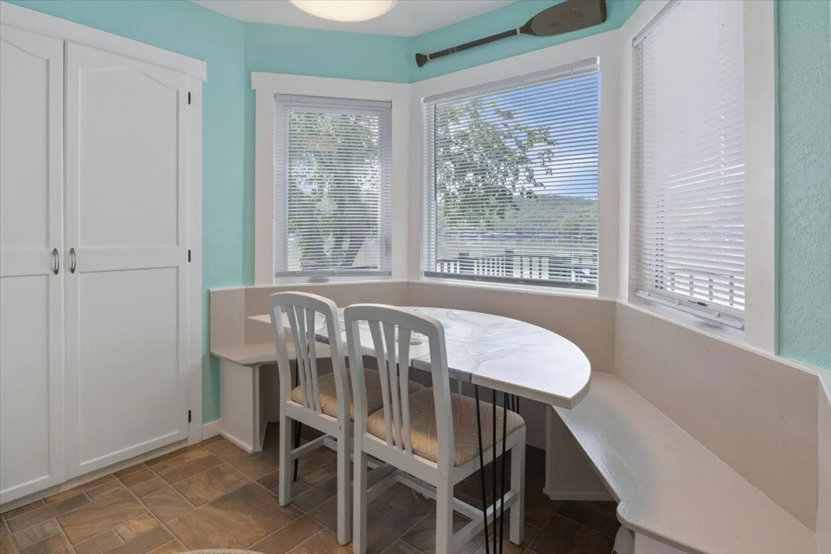 A curved dining table set beside large windows with white cabinetry nearby, allowing natural light to flood the space. The arrangement offers a scenic spot for group dining with views toward the outdoors.