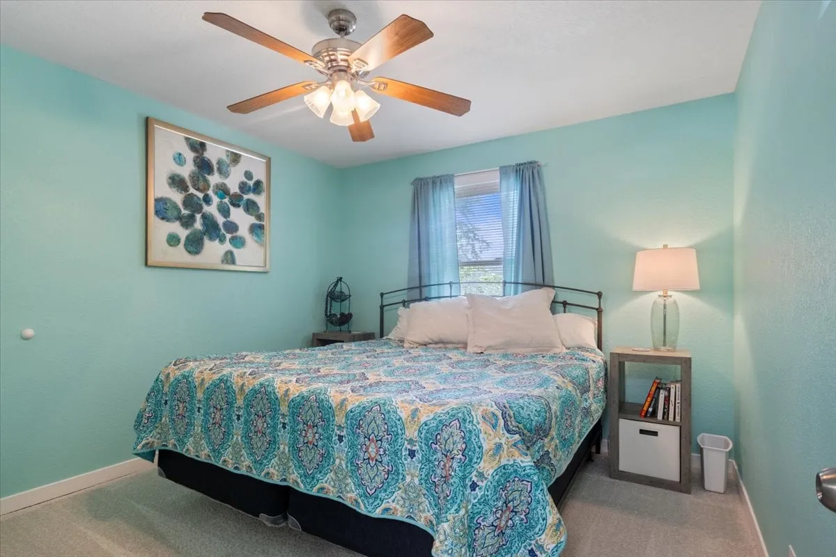 A cozy, tranquil bedroom featuring a bed with a patterned bedspread, a window with sheer curtains that lets in soft natural light, framed artwork on the wall, and a table lamp. The ambiance is peaceful and inviting, perfect for restful sleep or quiet reading moments.