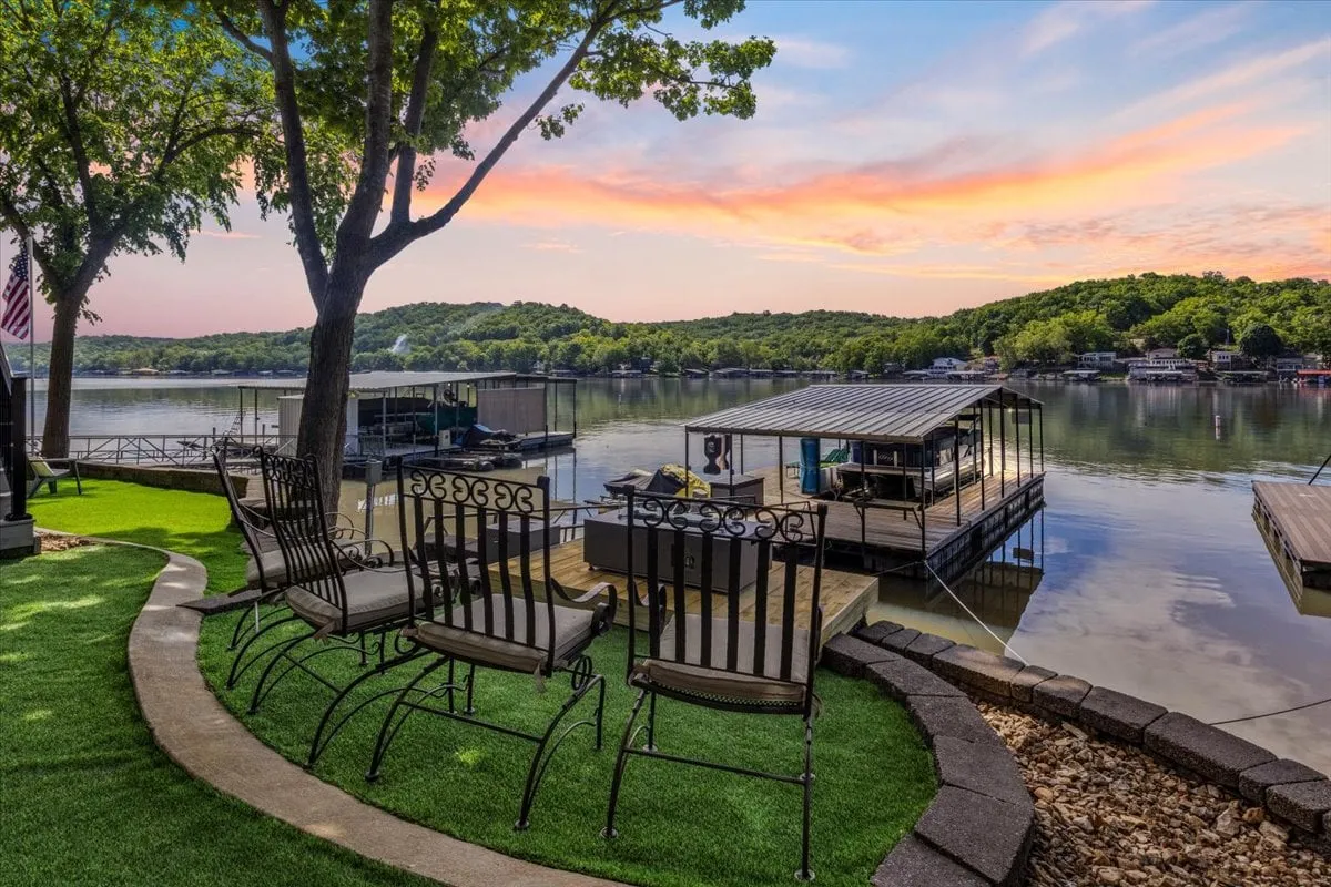 The image showcases a peaceful lakefront setting with a private dock extending into the sparkling waters of Lake of the Ozarks. Comfortable seating invites guests to relax under the shade of mature trees, while the fire pit and open sky hint at evenings spent sharing stories beside the water. The scene exudes tranquility, perfect for both quiet mornings with coffee and lively nights with friends.