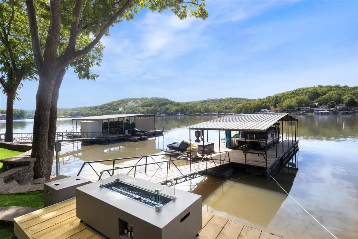 This exterior image features a serene lakeside scene with calm water, floating docks, and lush green hills with trees providing ample shade. The space invites various lakeside activities such as fishing, boating, relaxing, and gathering with friends or family, with a strong sense of tranquility and leisure.