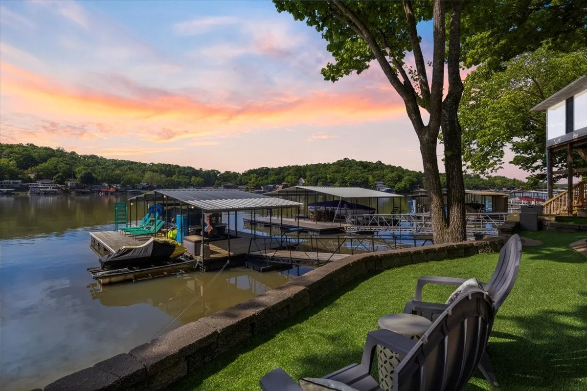 This exterior image shows the calm surface of the lake, several boathouses, a sunset sky, lush greenery, and an inviting outdoor seating area. The combination of natural beauty and inviting space highlights the appeal of mornings and evenings spent by the lake, whether for serene reflection or active water sports.