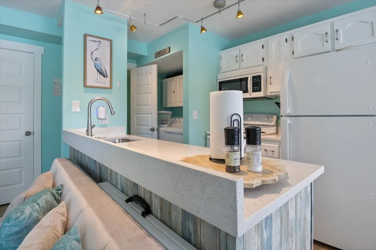 A bright, open kitchen featuring a central island perfect for both meal prep and socializing. Modern appliances line the countertops, and decorative artwork adds a cheerful touch. People are gathered, creating a lively and inviting atmosphere ideal for group cooking and entertaining.