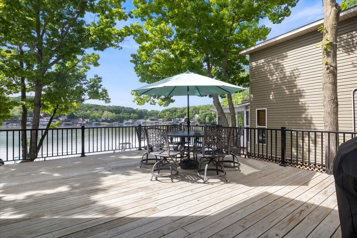 A spacious wooden deck set above the water, furnished with a large outdoor dining table and umbrella, surrounded by green trees and offering sweeping views of the lake. The setup invites guests to enjoy meals, morning coffee, or gatherings while soaking in the tranquil lakeside scenery.