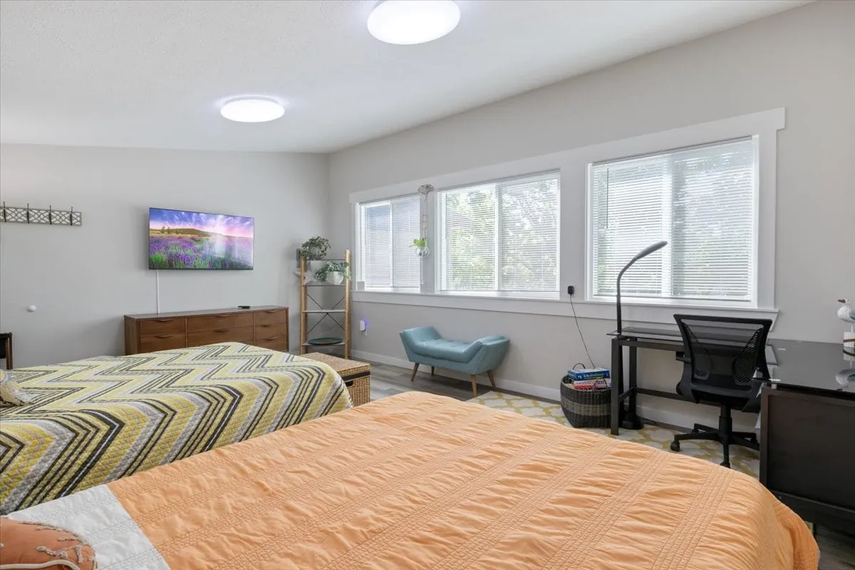 A comfortable bunkhouse-style room with beds adorned with patterned quilts, a wall-mounted television, large windows streaming in natural light, and a desk with a chair. The space is casual and versatile for relaxation, entertainment, or focused work.