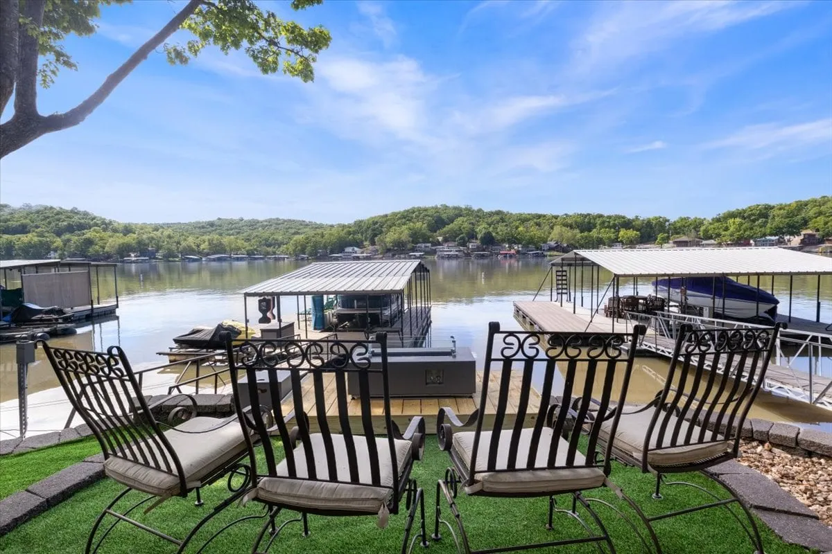This image captures a lakeside seating area with comfortable chairs overlooking the water and boathouses in the distance. The scene is calm, with the sun setting over the lake, and hints of a fire pit area, inviting guests to relax outdoors and enjoy evening conversations by the water.