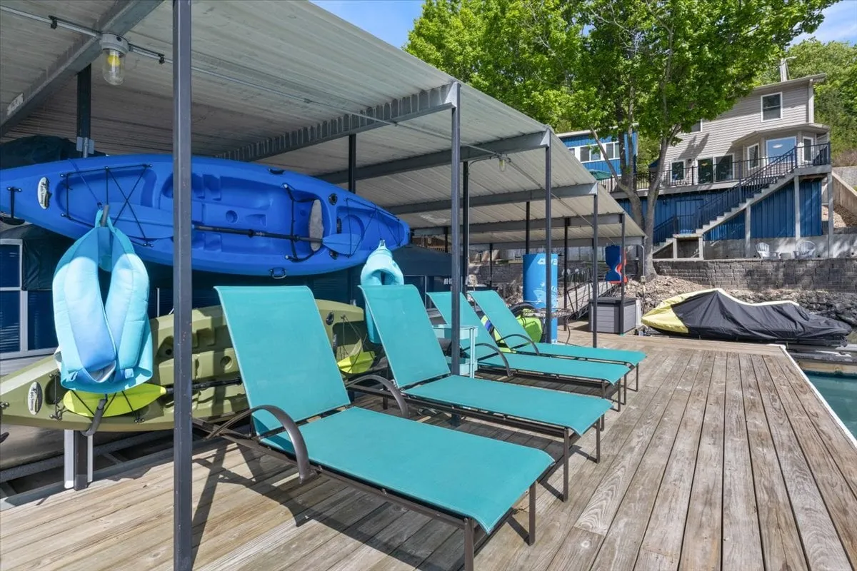 A sun-soaked wooden dock is the focal point, equipped with loungers, brightly colored kayaks, and life jackets. The setting overlooks calm lake waters, inviting guests to embark on kayaking adventures or relax in the sun.