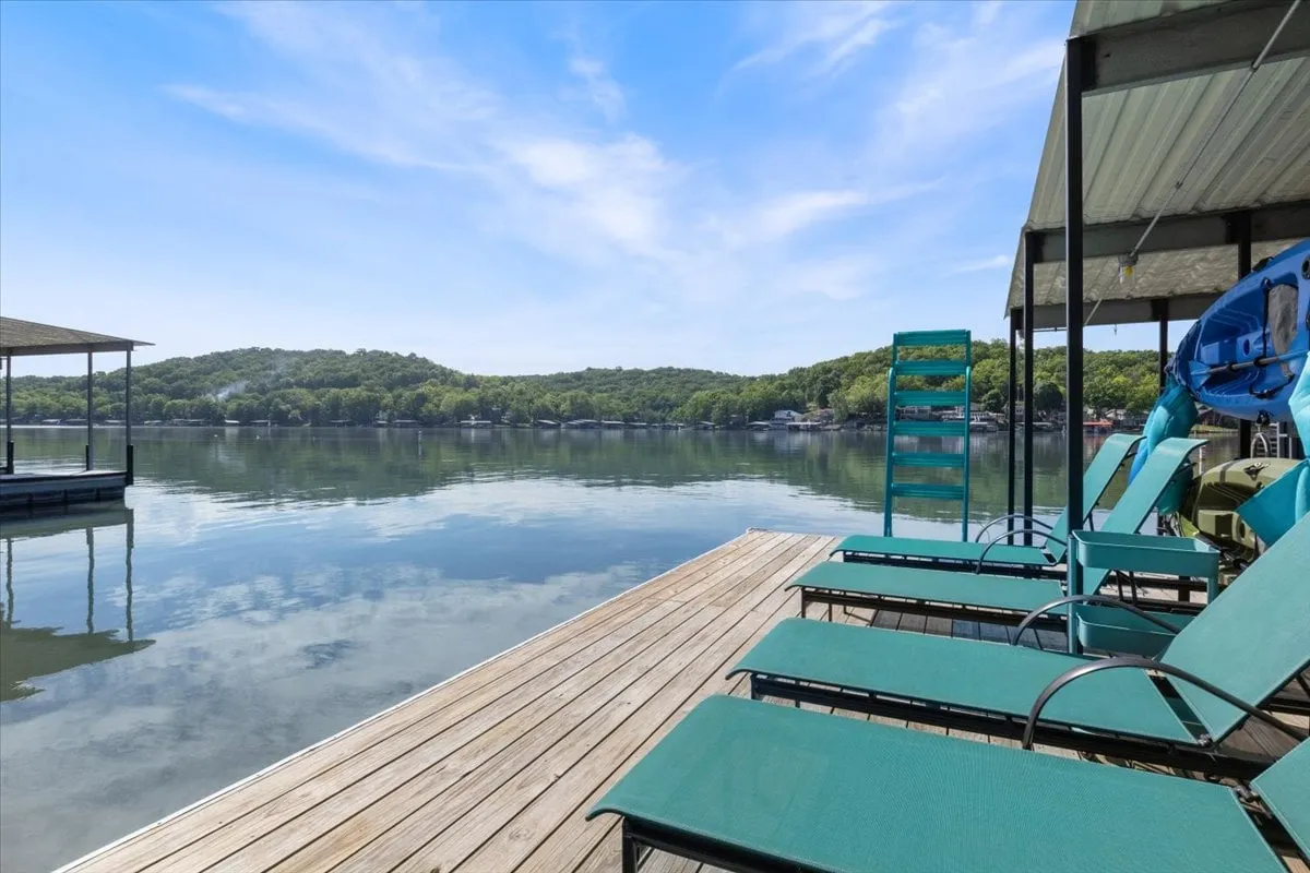 A wooden dock extends onto the calm water, flanked by lounge chairs and a boathouse. The green hillside and tranquil lake create the ideal spot for sunbathing, social gatherings, or launching a kayak at sunset.